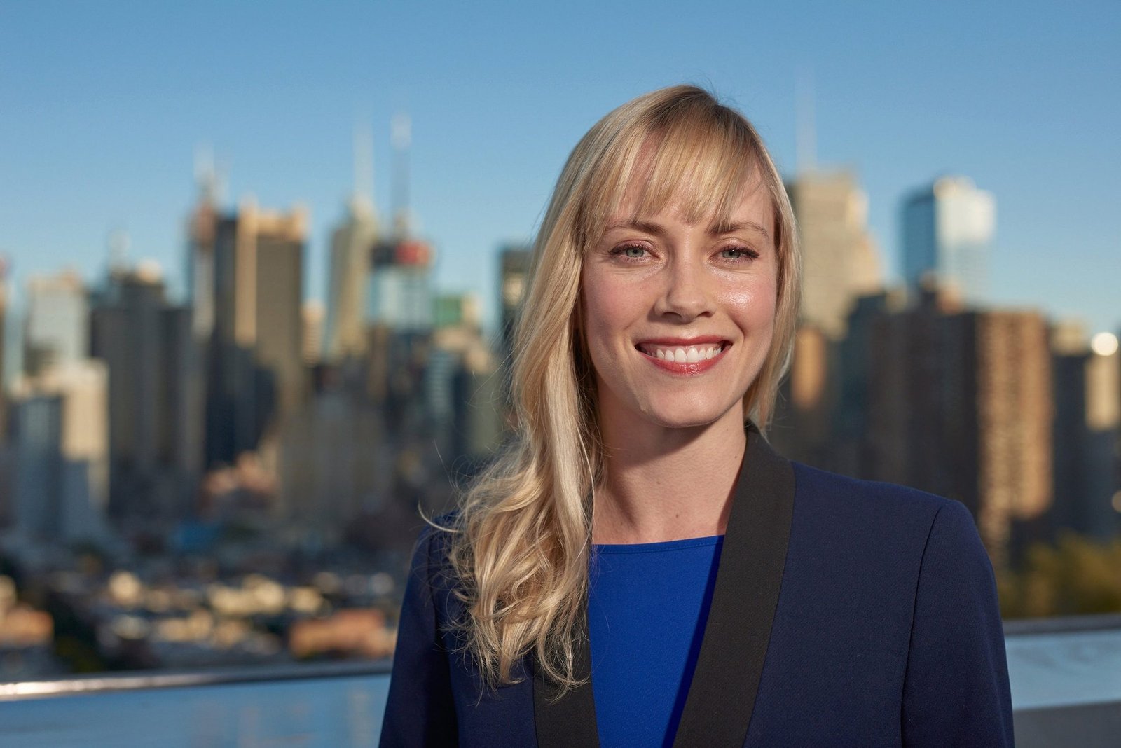 Corporate headshot of a smiling woman in a blue blazer with a city skyline behind her