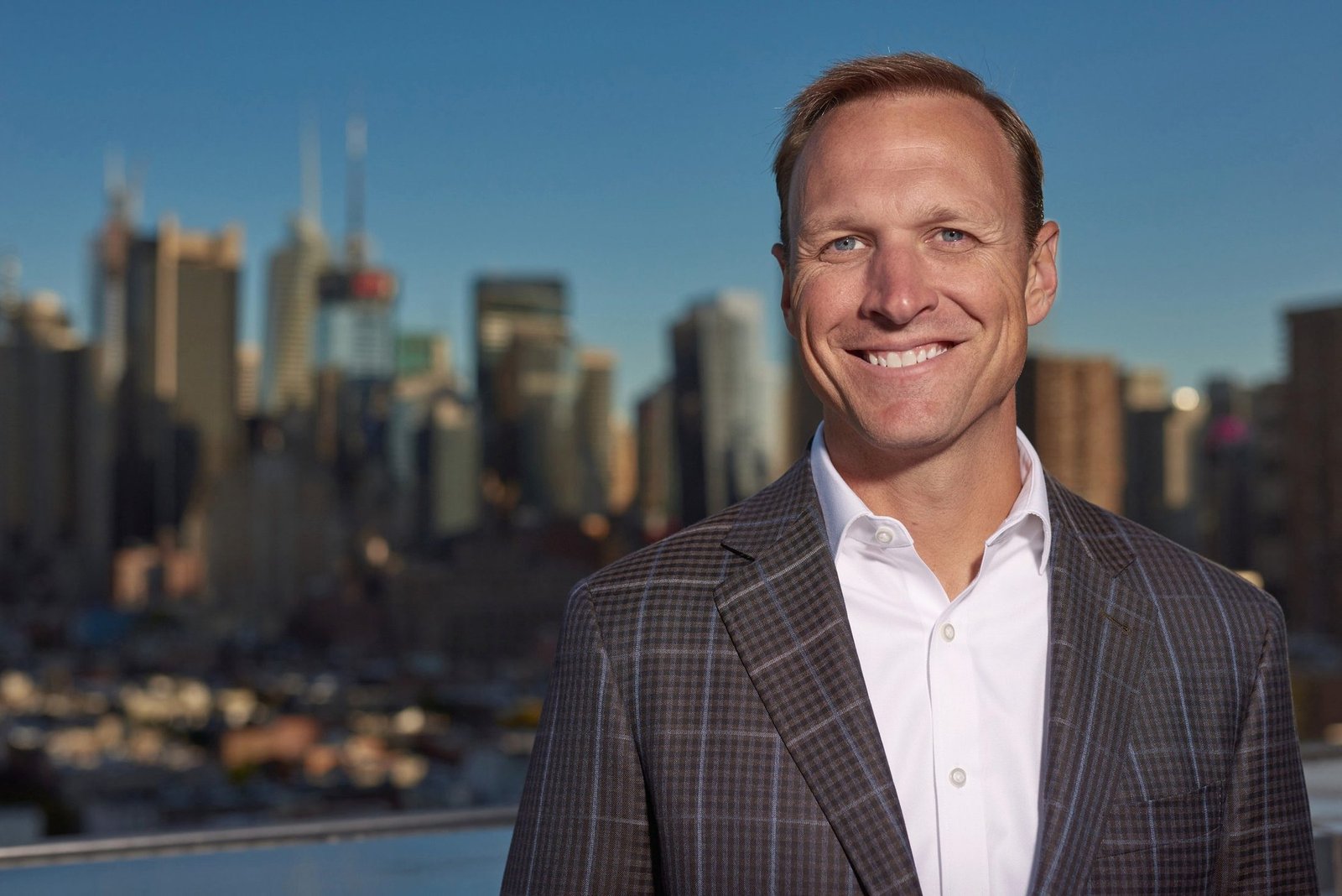 Executive headshot of a man in a checkered blazer on a rooftop with a city skyline