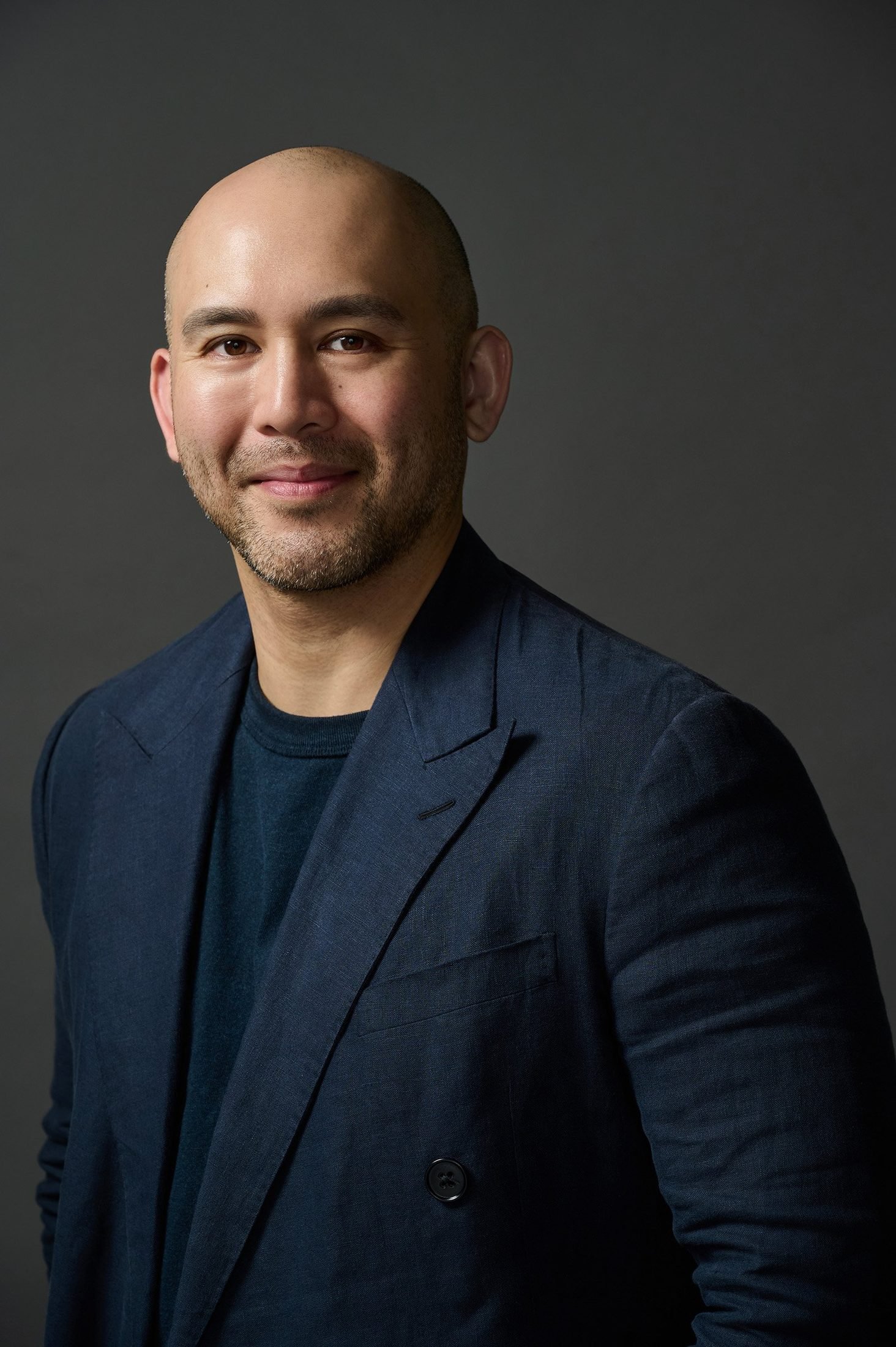 Studio headshot of a man with a shaved head and beard in a dark blazer